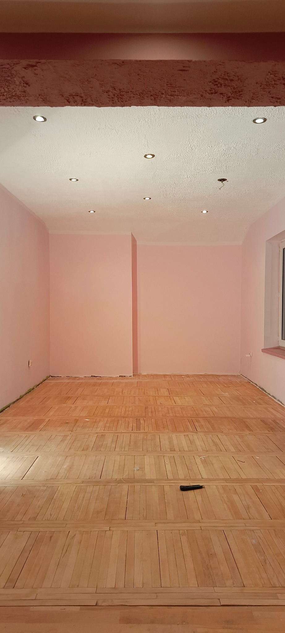 Pokój z różowymi ścianami i częściowo ułożoną podłogą z drewnianych paneli; widoczny czarny nóż do tapet na podłodze.