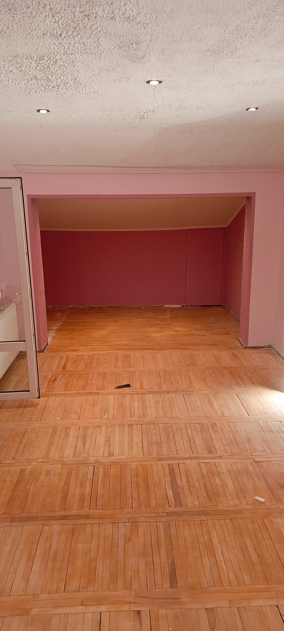Wnętrze pokoju z ułożoną drewnianą podłogą z listew, różowe ściany i sufit z oświetleniem punktowym, widoczny fragment listwy przypodłogowej i mały czarny przedmiot na podłodze.