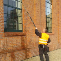 Pracownik w kamizelce odblaskowej czyści okno na wysokości za pomocą teleskopowej szczotki na tle ceglanej elewacji.