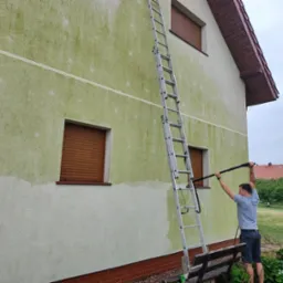 Pracownik w Wałbrzychu czyści elewację domu z zielonego nalotu za pomocą lancy ciśnieniowej, stojąc na drabinie przystawionej do ściany.