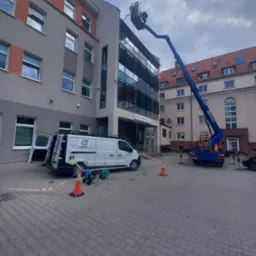 Pracownik w żółtym kombinezonie na podnośniku koszowym, przy elewacji budynku z licznymi oknami; biały van firmy Osmotic z otwartymi drzwiami, zabezpieczenia w postaci taśmy i pachołków...