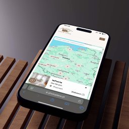 SOCIALBUZZSTUDIO Sp. z o.o. - Ekran smartfona z otwartą aplikacją Photo Place, prezentującą mapę Polski z lokalizacjami i cenami usług fotograficznych. Telefon leży na drewnianej powierzchni.