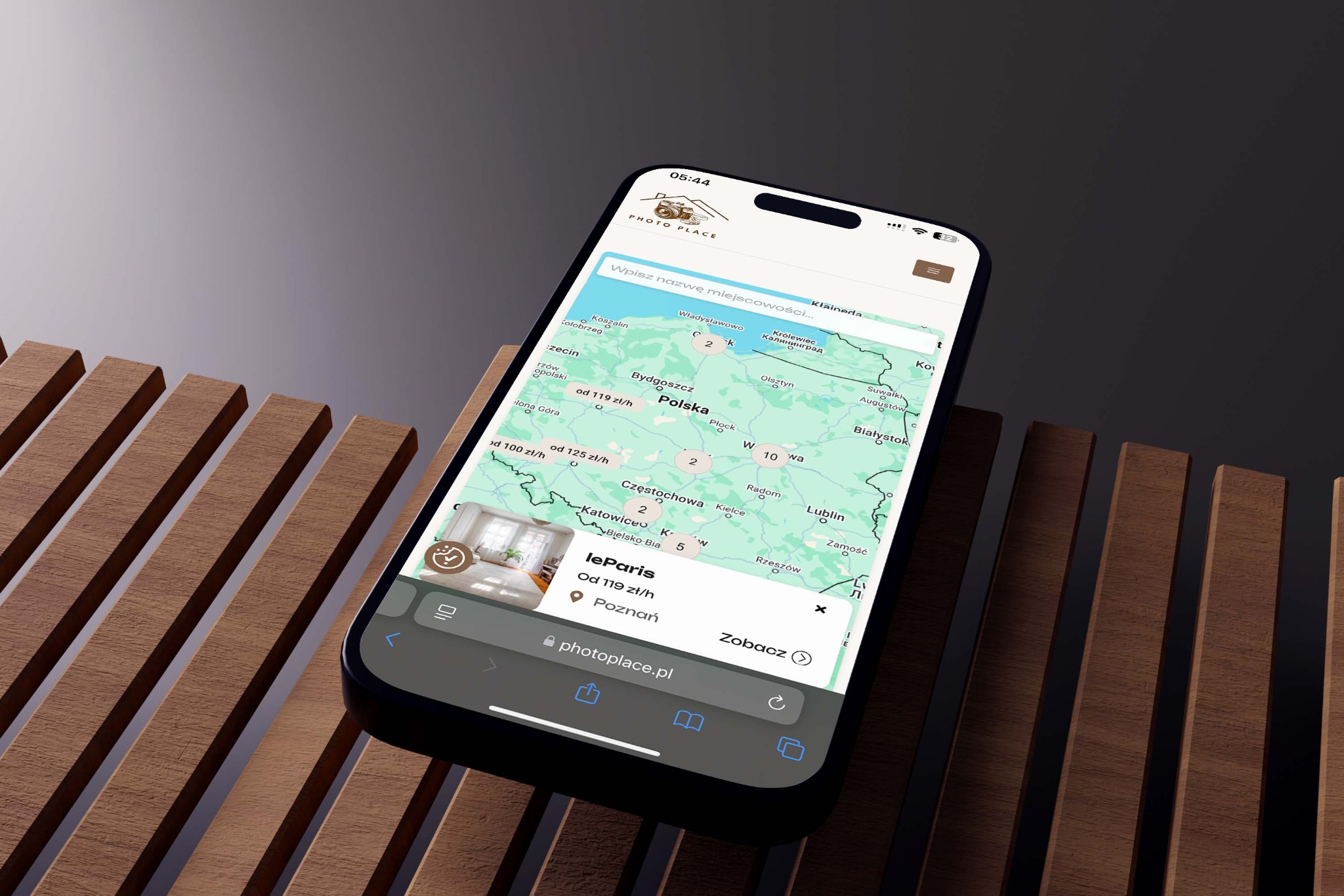 Ekran smartfona z otwartą aplikacją Photo Place, prezentującą mapę Polski z lokalizacjami i cenami usług fotograficznych. Telefon leży na drewnianej powierzchni.