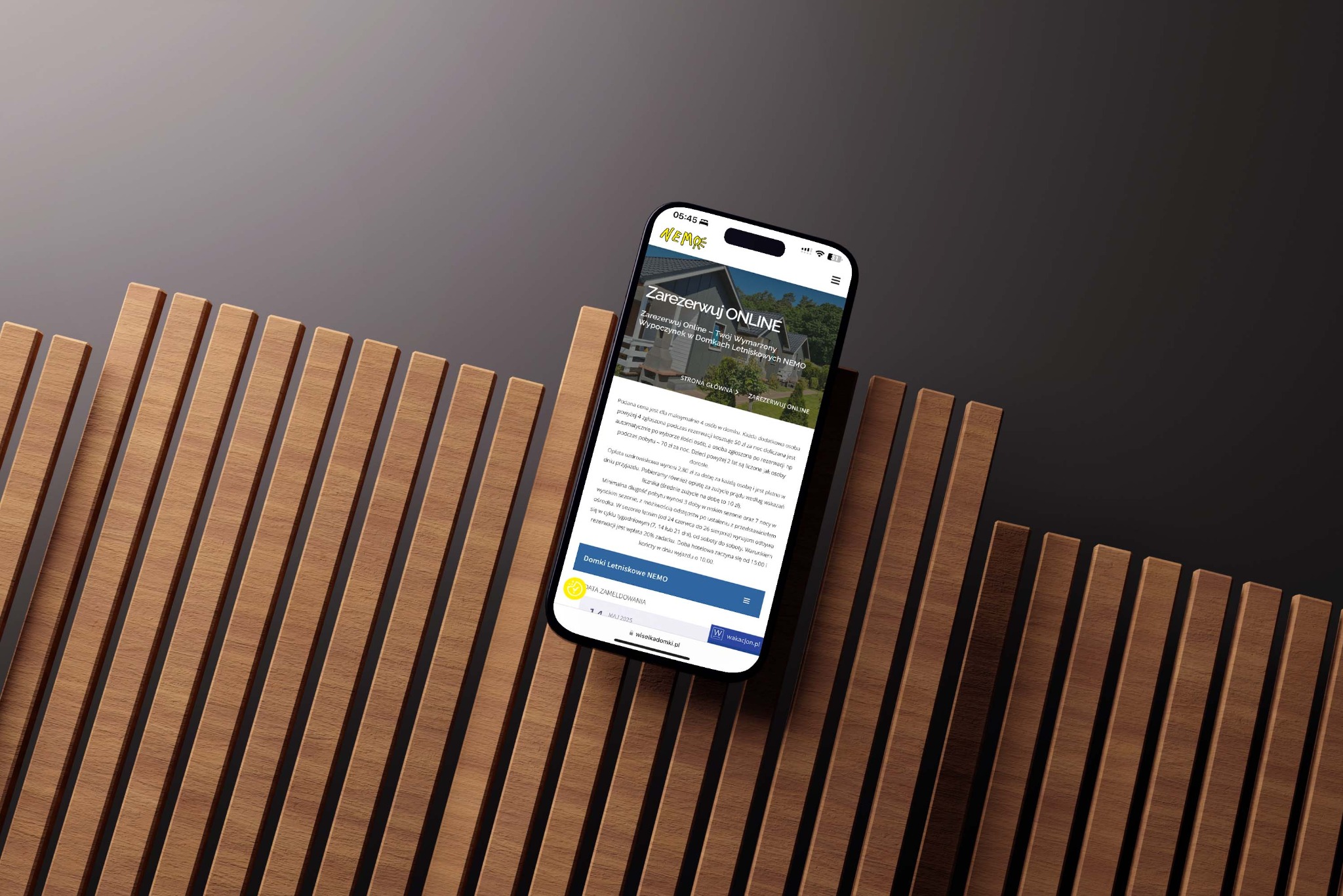 Smartfon prezentuje stronę internetową z możliwością rezerwacji domków letniskowych NEMO, umieszczony na tle z drewnianych listew.