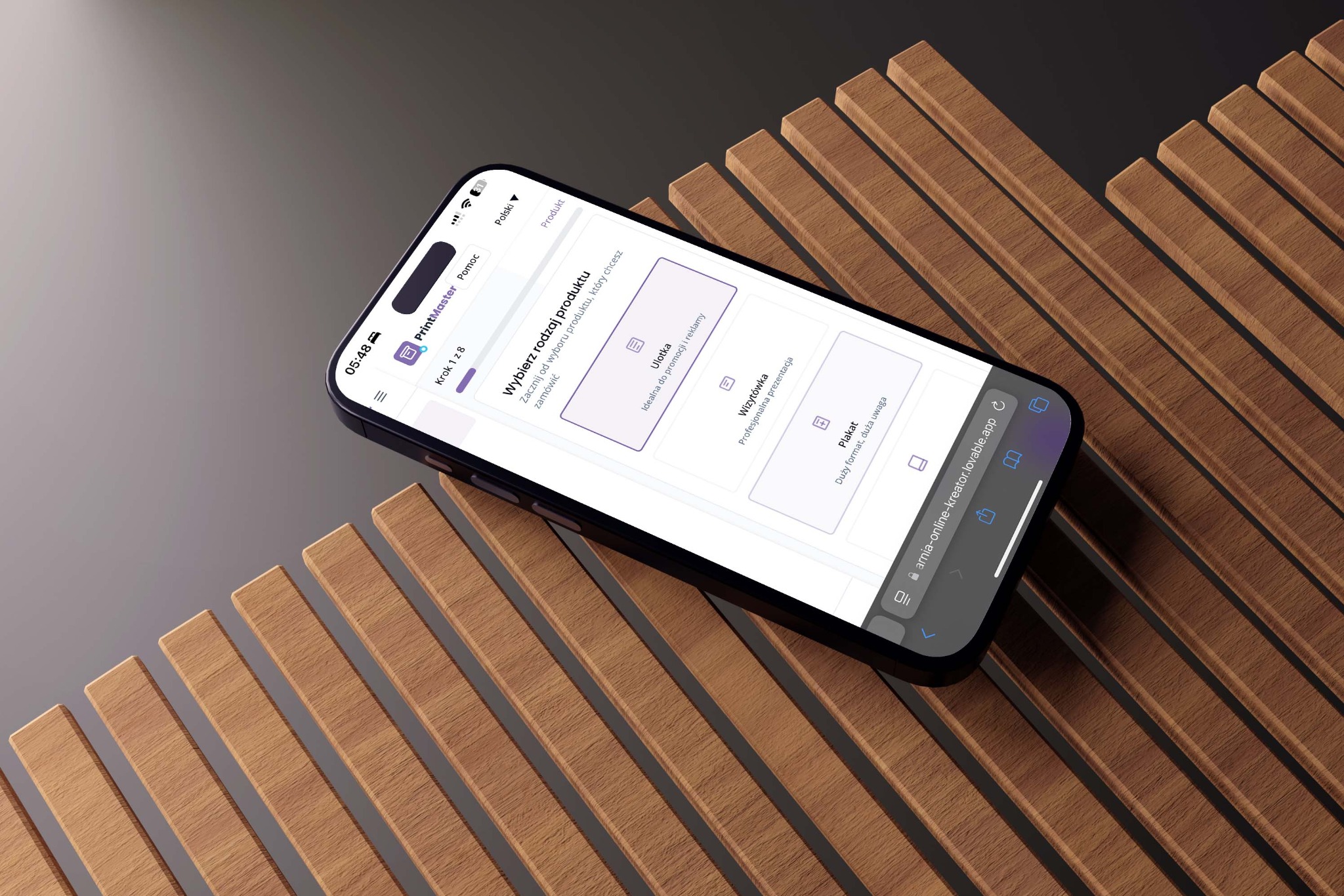 Aplikacja PrintMaster na smartfonie, prezentująca opcje wyboru produktu do druku: ulotki, wizytówki, plakaty. Telefon leży na drewnianym tle z listewek.