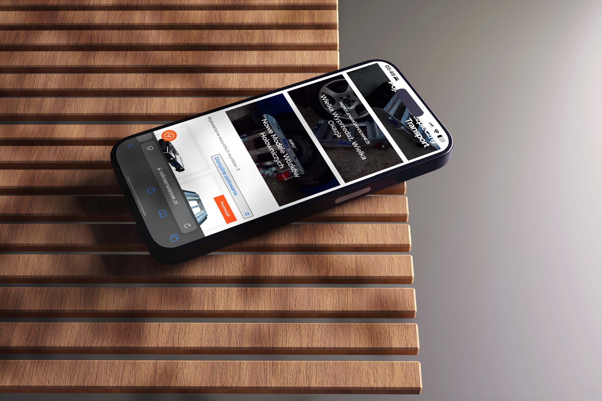 Smartfon wyświetlający stronę internetową z ofertami wózków holowniczych, leżący na drewnianym tle. Widoczny pasek adresu i przyciski nawigacyjne przeglądarki.