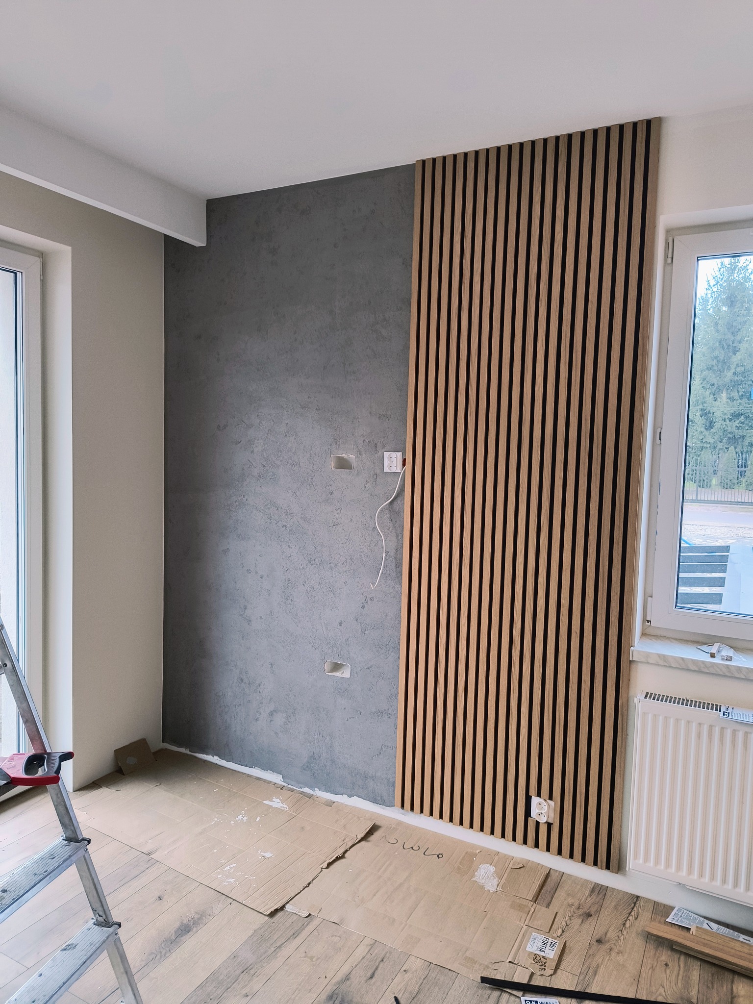 Szara ściana z fakturą betonu sąsiaduje z drewnianymi lamelami na fragmencie ściany, widoczne gniazdka elektryczne, podłoga zabezpieczona kartonem, fragment drabiny malarskiej.