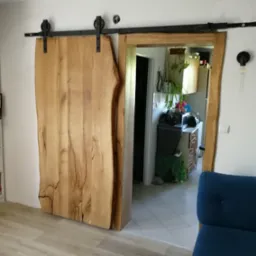 Drewniane drzwi przesuwne z surowego drewna na czarnych szynach, widok na kuchnię w tle.