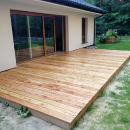 Nowoczesny taras drewniany z jasnych desek, przylegający do domu z dużymi przesuwnymi drzwiami tarasowymi, widok na zieleń w tle.