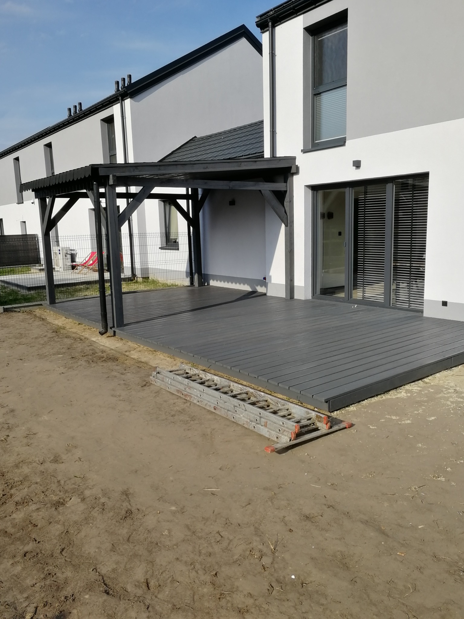 Szary taras z drewnianych desek przylegający do nowoczesnego domu z czarną pergolą, na ziemi leżąca drabina.