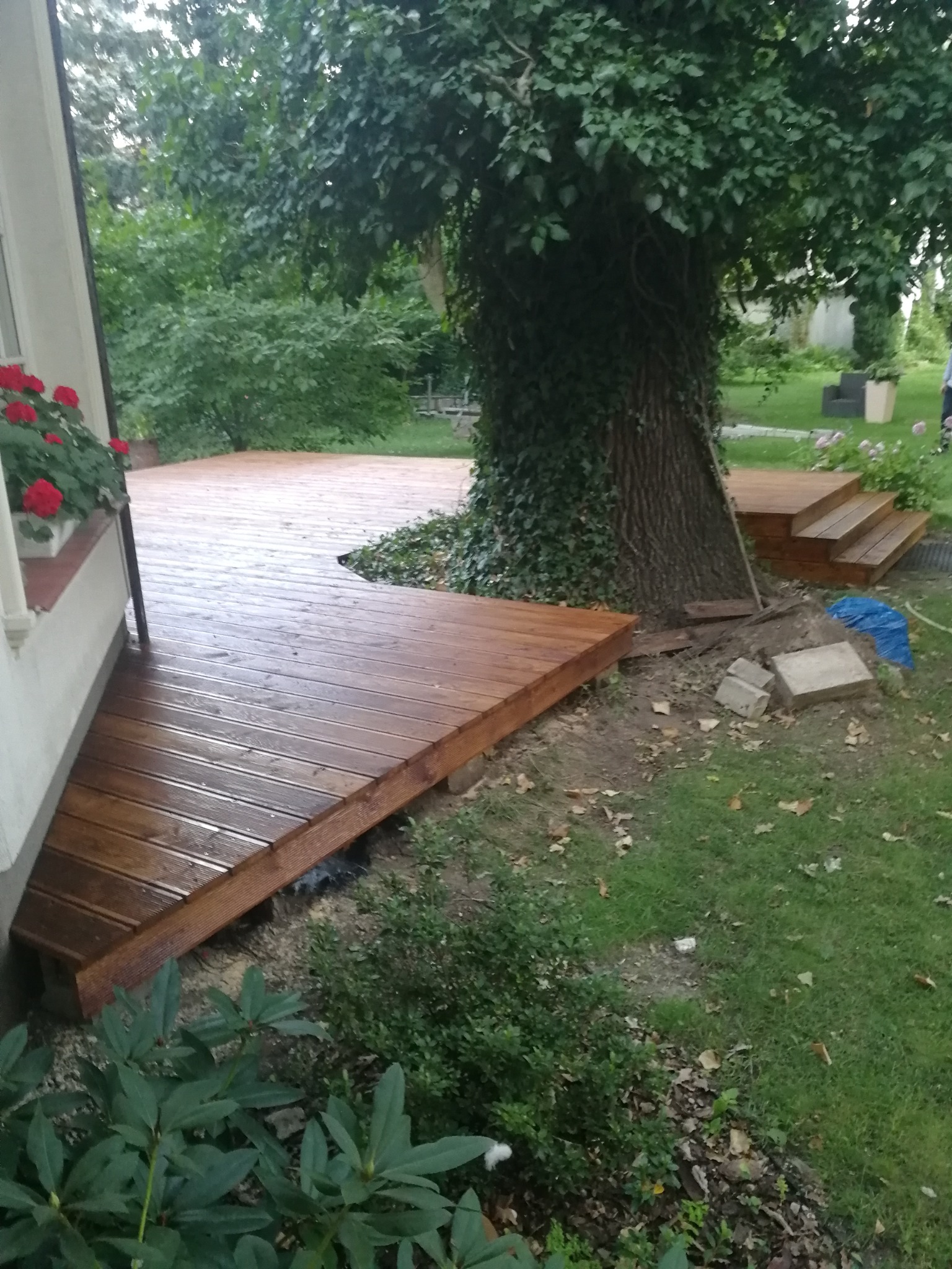 Brązowy taras przylegający do domu, z czerwonymi kwiatami w oknie, otoczony zielenią ogrodu i dużym drzewem z bluszczem.