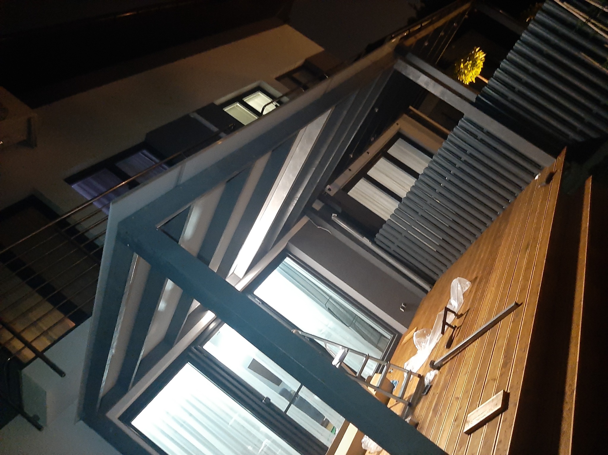 Montaż konstrukcji z metalowymi ramami i drewnianym wypełnieniem, widoczne rusztowanie i zabezpieczenia, ujęcie z dołu w warunkach słabego oświetlenia.