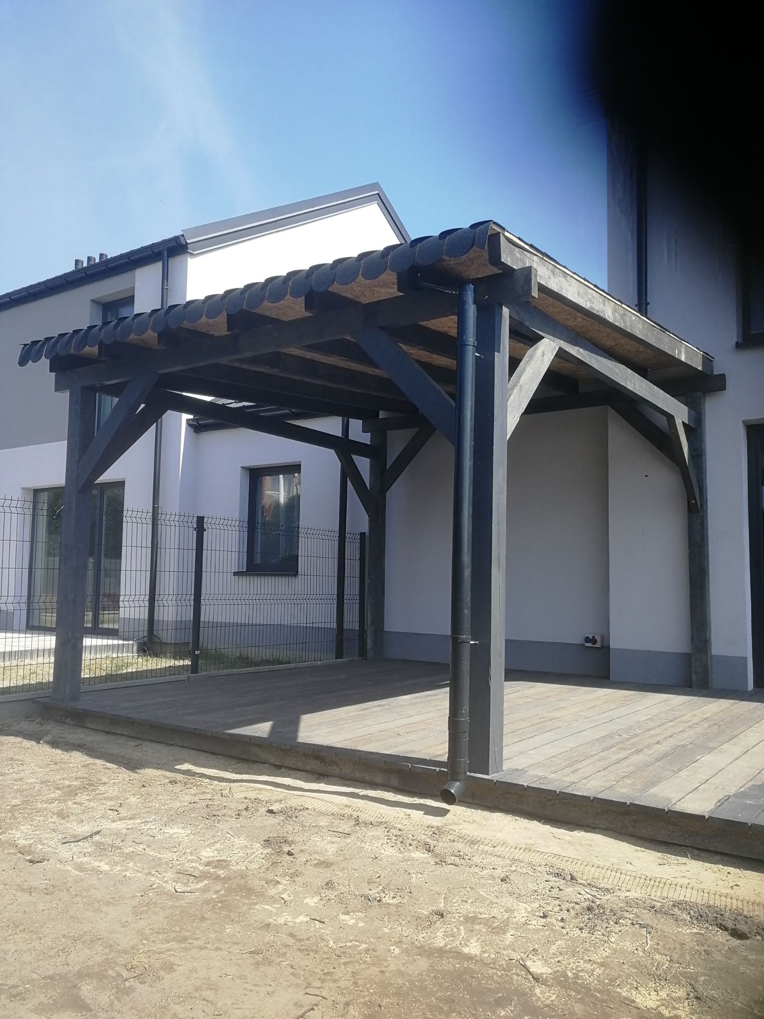 Ciemna drewniana konstrukcja z zadaszeniem na podwyższonym drewnianym tarasie, widok z zewnątrz na tle nowoczesnego domu jednorodzinnego i ogrodzenia.