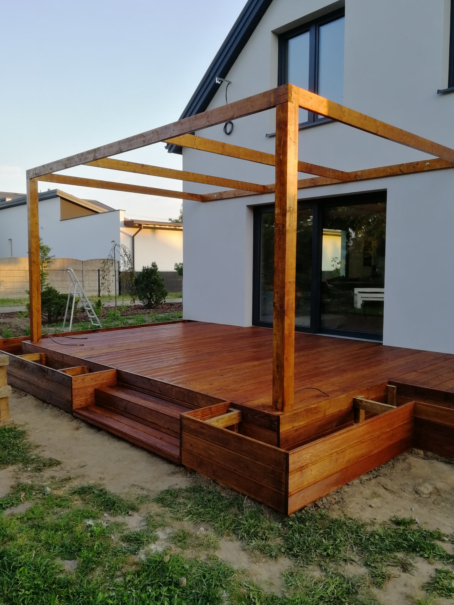 Nowoczesny taras z drewna egzotycznego w trakcie budowy, widoczne schody, podwyższone rabaty i konstrukcja przyszłej pergoli, w tle biały dom z dużymi oknami przesuwnymi.