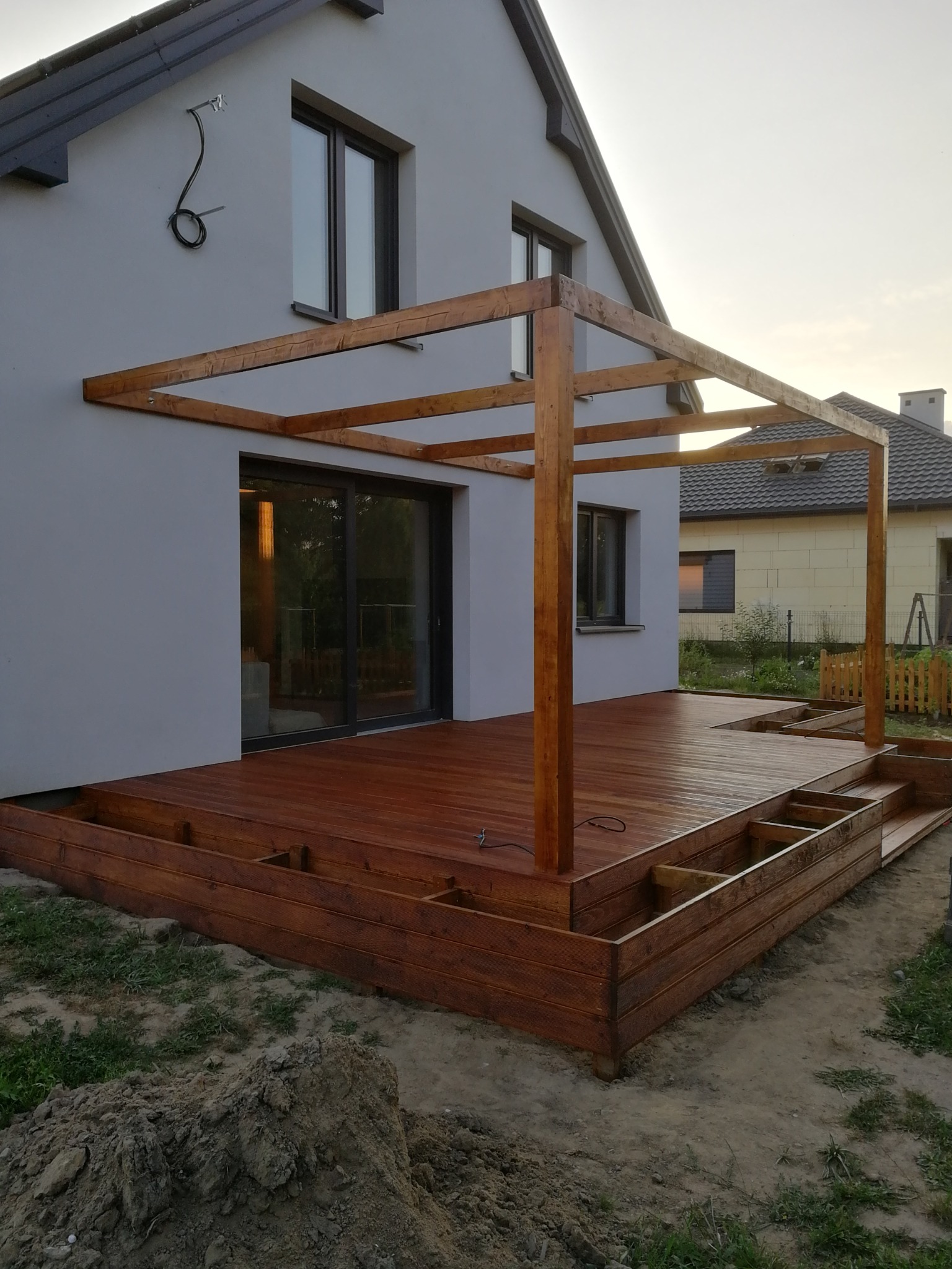 Drewniany taras z pergolą w trakcie budowy, widoczne elementy konstrukcyjne i fragmenty desek, dom z szarą elewacją w tle.
