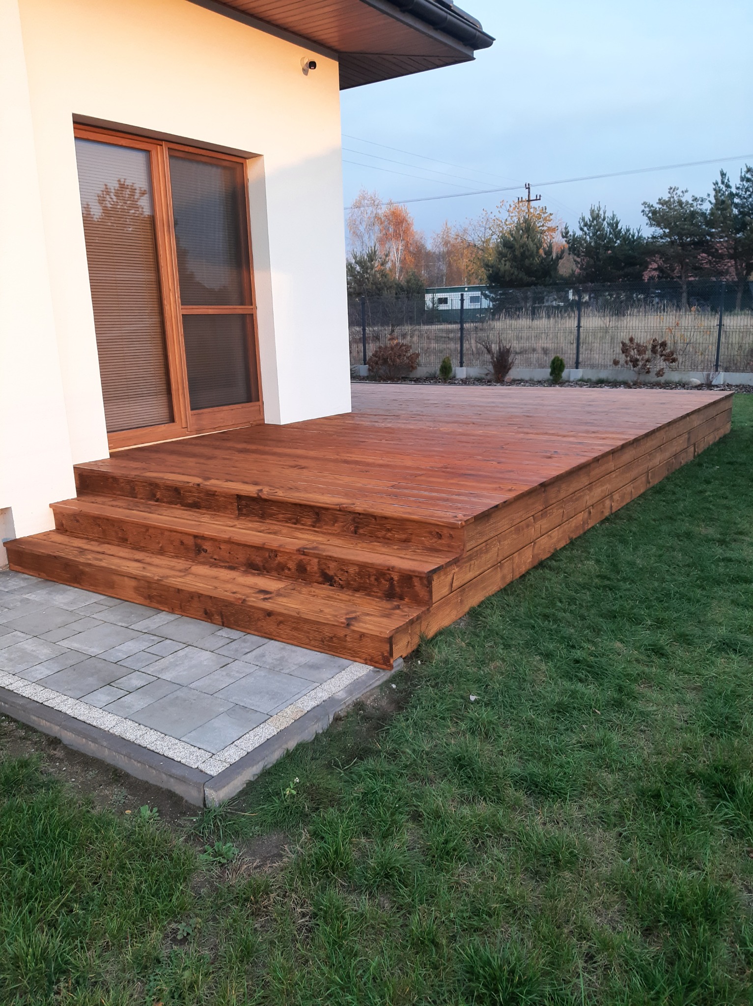 Nowoczesny taras z drewna, z trzema stopniami prowadzącymi do drzwi balkonowych, wykończony kostką brukową i otoczony trawnikiem.