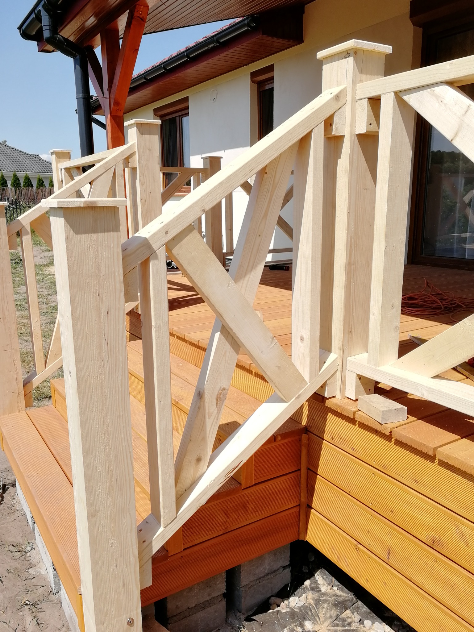 W trakcie budowy: drewniany taras z surową, jasną balustradą i widocznymi elementami konstrukcyjnymi, częściowo pomalowany na brązowo, przylegający do białego domu z brązowymi oknami.