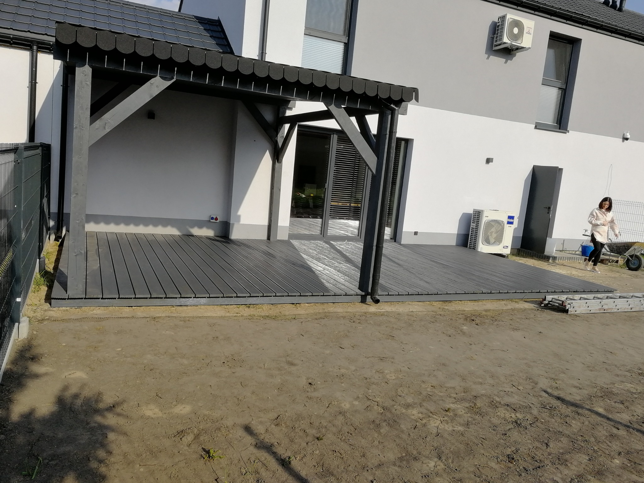 Nowoczesny, szary taras z drewnianą pergolą przy domu jednorodzinnym, widoczna drabina i osoba w tle, teren wokół w trakcie prac ziemnych.