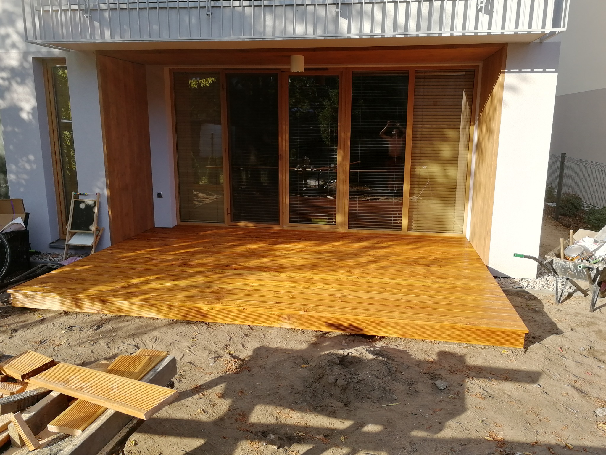 Nowo wybudowany, drewniany podest przy domu z widocznymi deskami do wykończenia, taczką i narzędziami na zewnątrz.