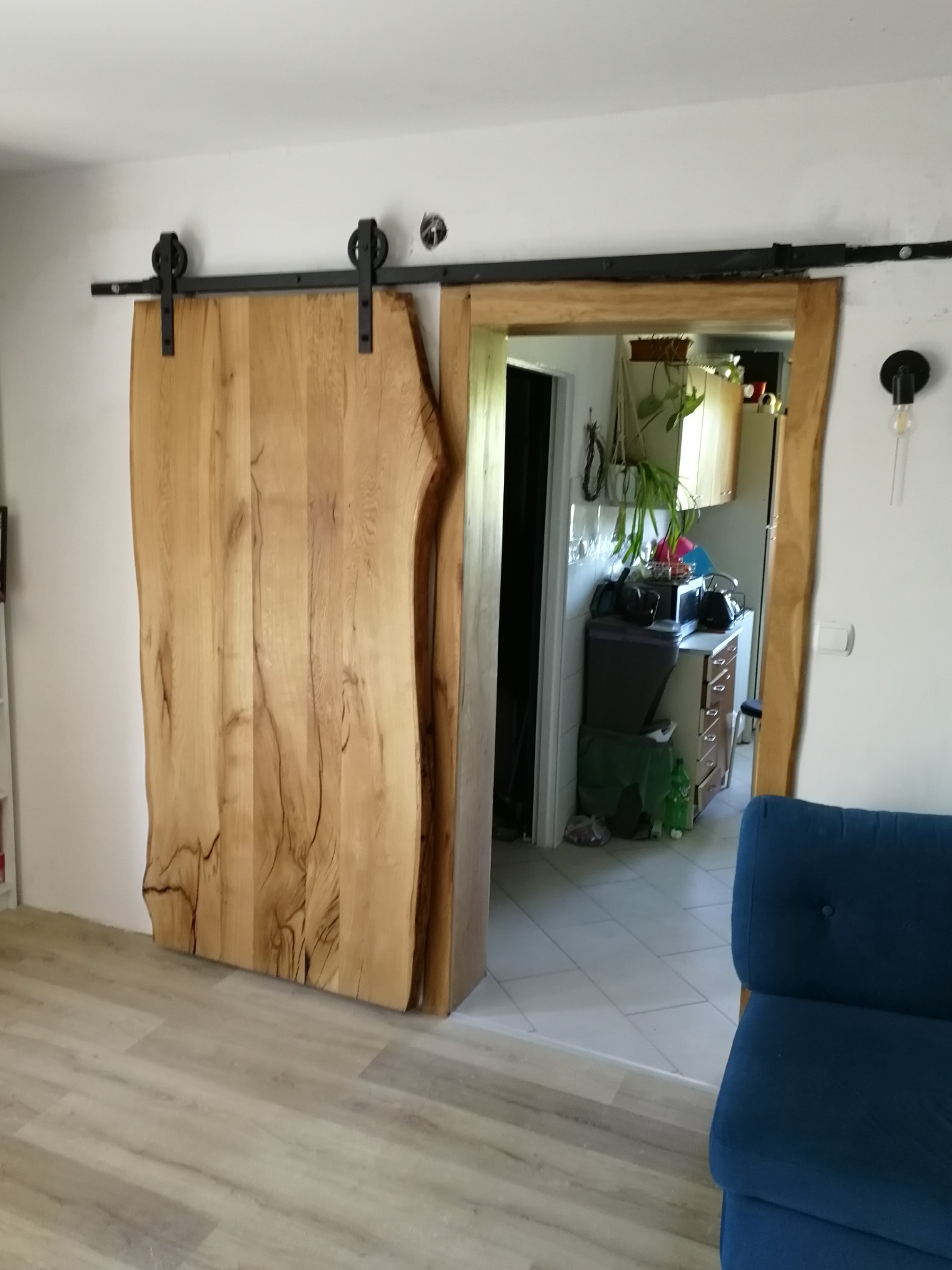 Drewniane drzwi przesuwne z surowego drewna na czarnych szynach, widok na kuchnię w tle.