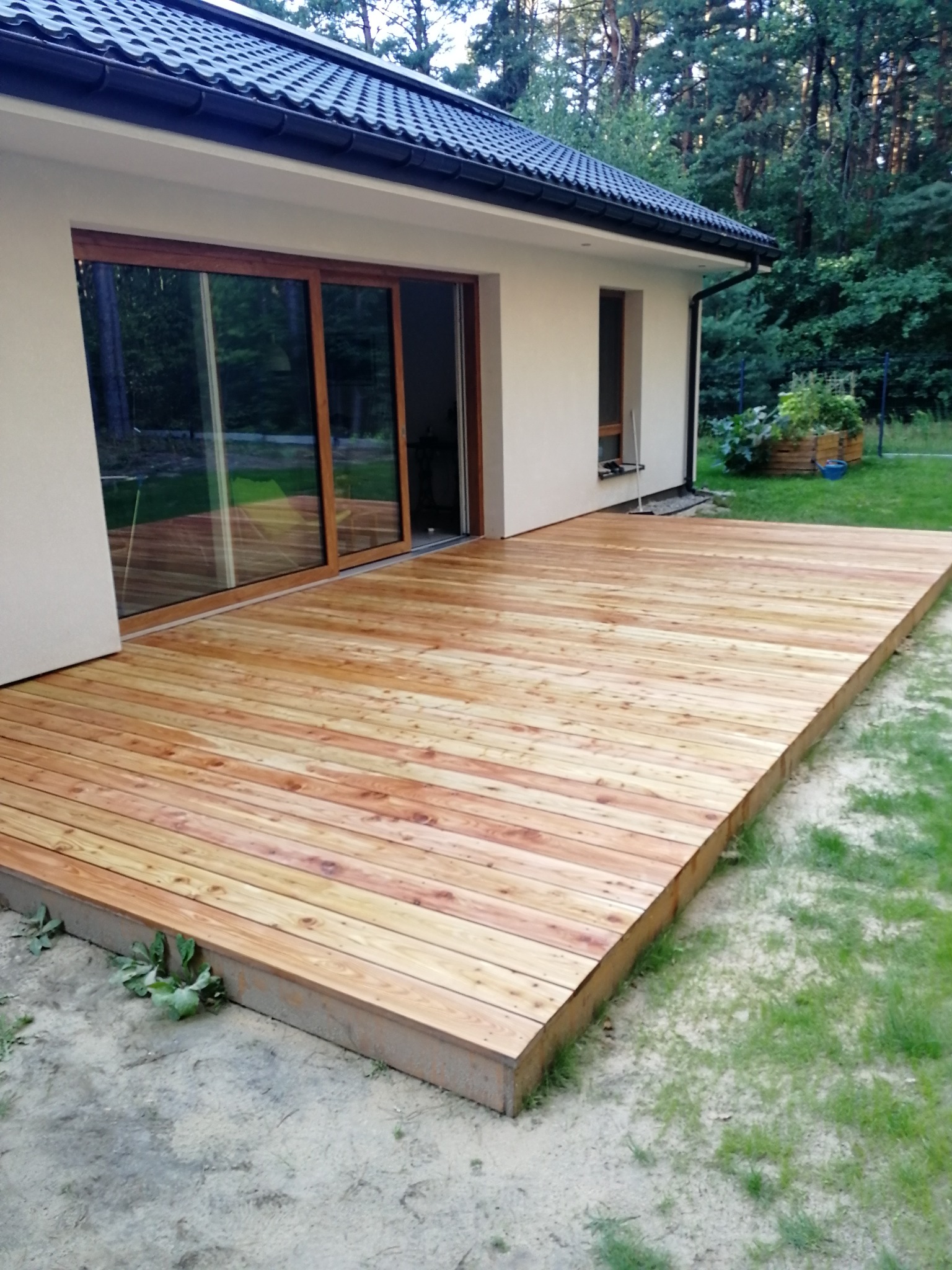 Nowoczesny taras drewniany z jasnych desek, przylegający do domu z dużymi przesuwnymi drzwiami tarasowymi, widok na zieleń w tle.