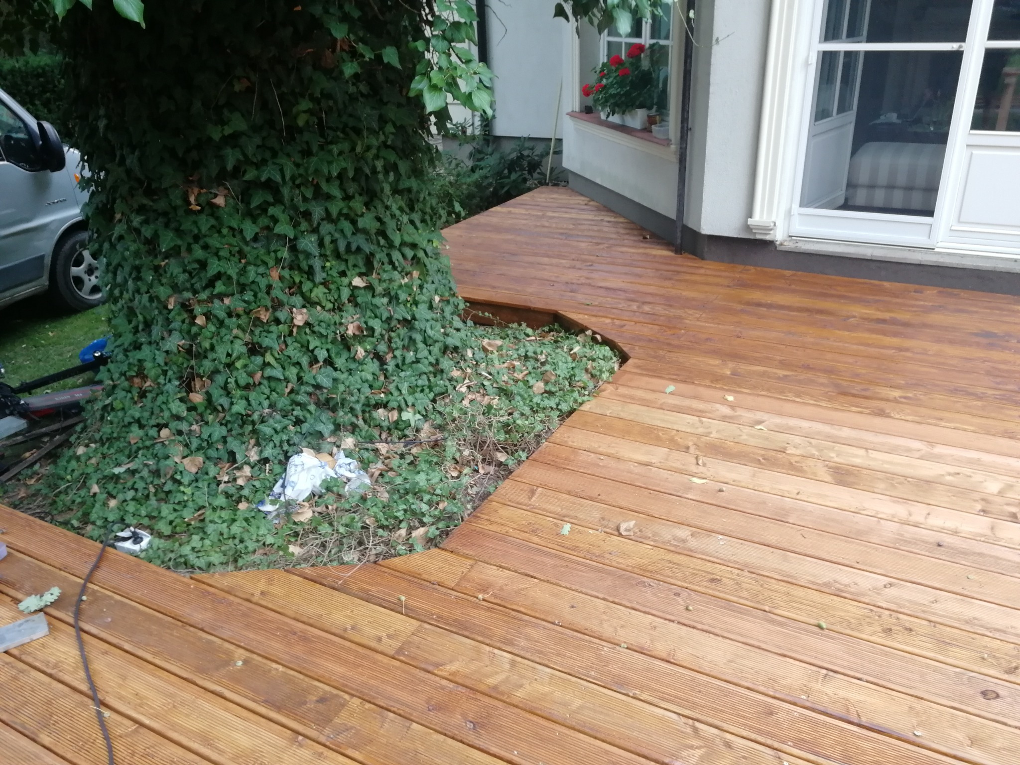 Nowo wykonany taras z desek wokół pnia drzewa, z widocznym fragmentem samochodu i domu w tle.