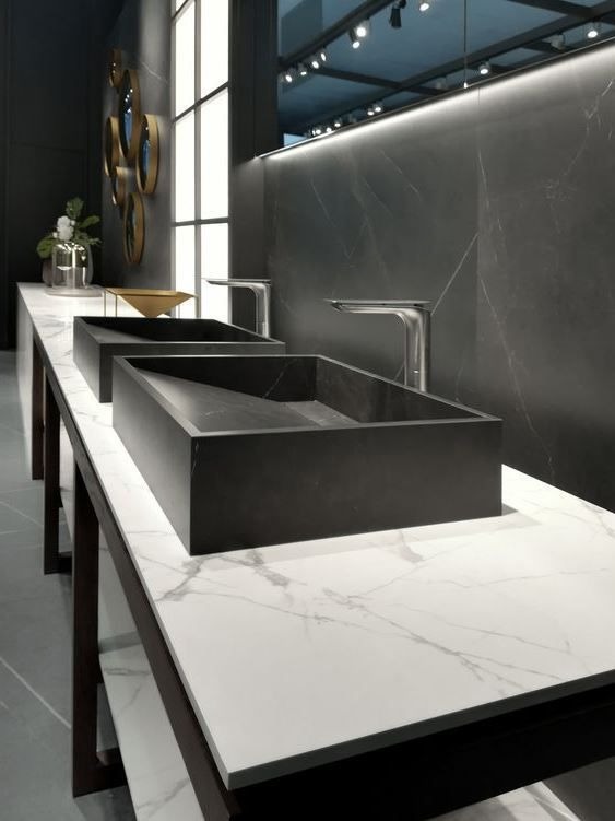 Dwie prostokątne, czarne umywalki nablatowe z chromowanymi bateriami na blacie z białego marmuru z szarymi żyłami, na tle ściany z ciemnoszarego marmuru i okrągłych dekoracji ściennych w odcieniach...