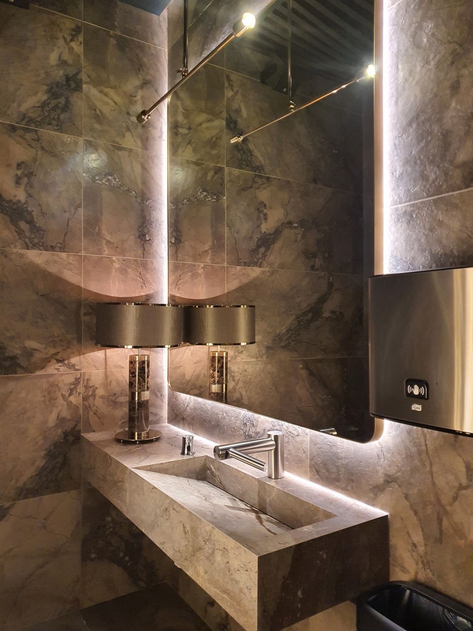 Nowoczesna umywalka z szarego marmuru z wbudowanym oświetleniem LED, uzupełniona chromowaną armaturą i dwoma lampami stołowymi z abażurami, na tle ściany wyłożonej płytkami imitującymi marmur.