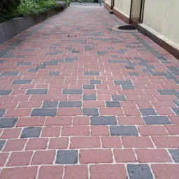 Ułożona kostka brukowa w odcieniach czerwieni i szarości, tworząca wzór. Widok na czystą, zadbaną nawierzchnię chodnika przy budynku i zieleni.