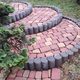 Marcin Stulfig PMC clean bruk - Kręta ścieżka z czerwonej kostki brukowej obrzeżona szarymi palisadami, wijąca się przez ogród z zielonymi krzewami i drzewami iglastymi w tle.