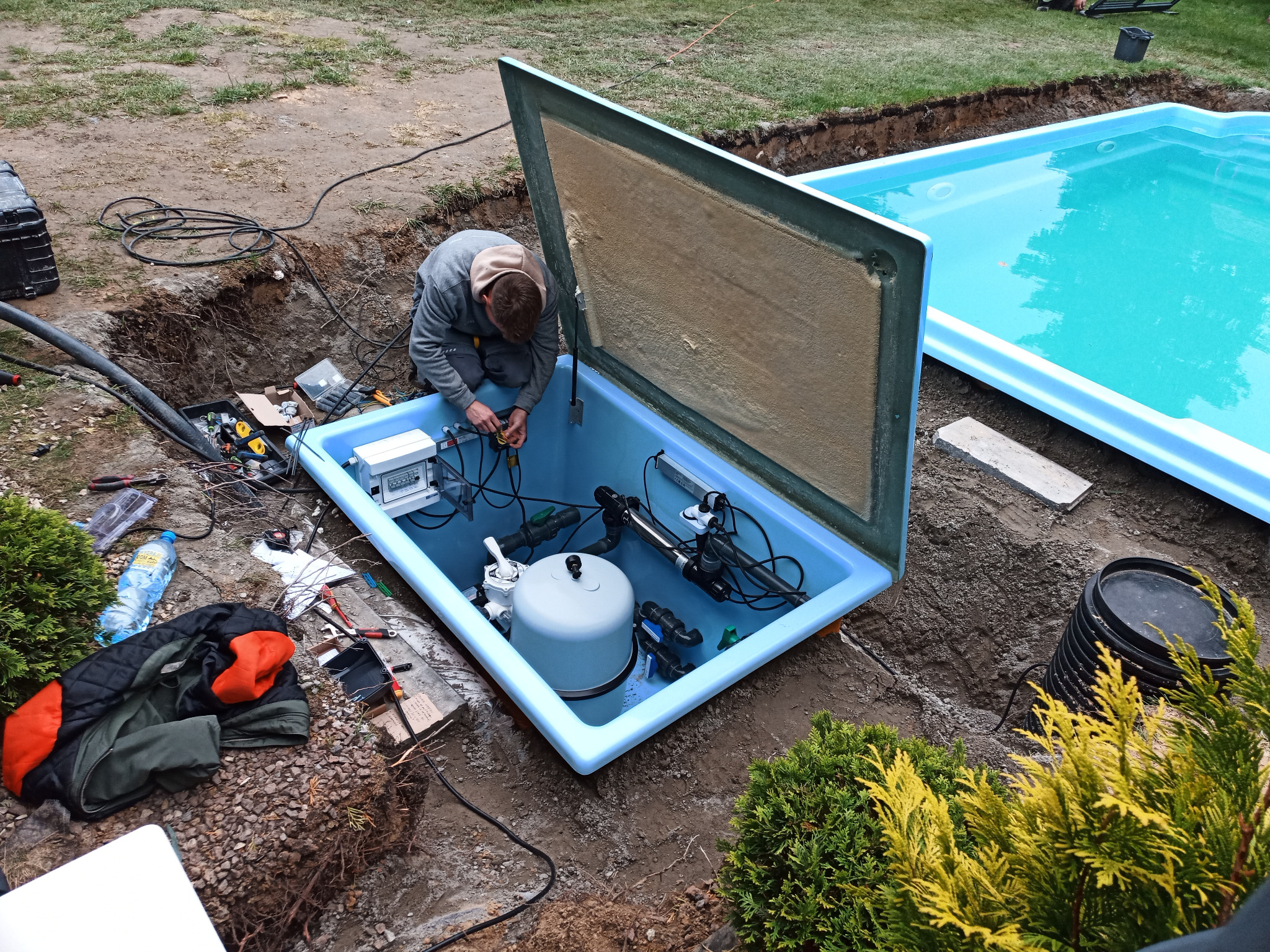 Instalacja podziemnej skrzynki z osprzętem do basenu, otwartej, z widocznymi filtrami i pompami, technik podłącza okablowanie, obok basen z wodą.