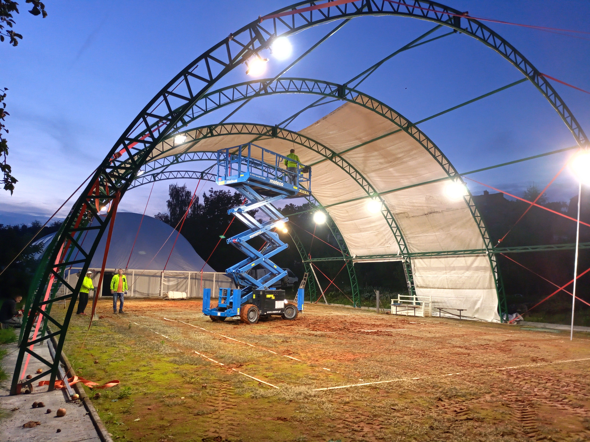 Montaż oświetlenia hali sportowej z użyciem podnośnika koszowego, widoczne stalowe łuki konstrukcji i pracownicy w odblaskowych kamizelkach.