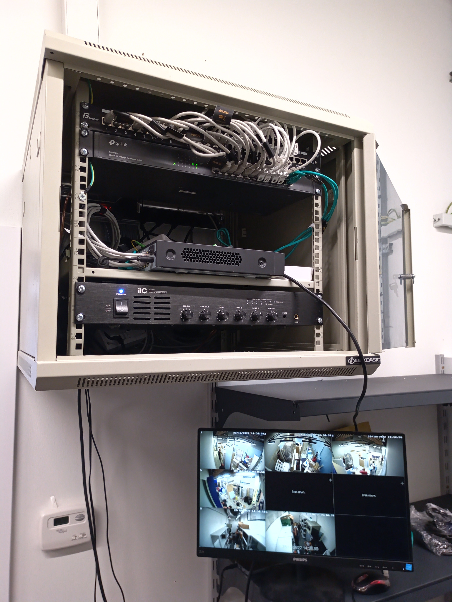 Szafa rack z zainstalowanymi urządzeniami sieciowymi i mikserem audio, poniżej monitor z podglądem z kamer monitoringu.