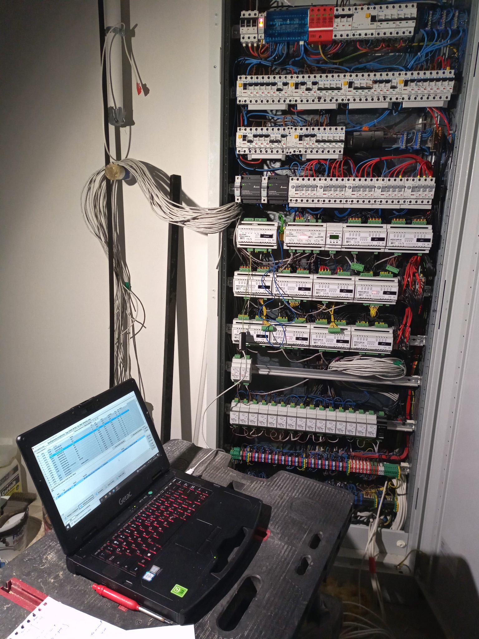 Otwarta szafa sterownicza z licznymi przewodami i zabezpieczeniami, laptop Getac na stołku roboczym z widocznym oprogramowaniem do konfiguracji, kable zwisające ze ściany.
