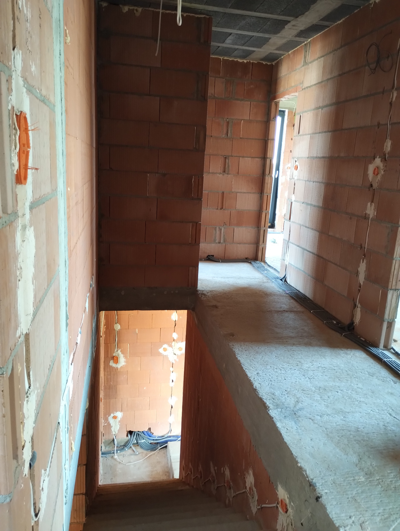 Wnętrze budynku w trakcie budowy, widoczne instalacje elektryczne w ścianach z cegły, korytarz z betonową wylewką i strop z szarej wełny mineralnej.