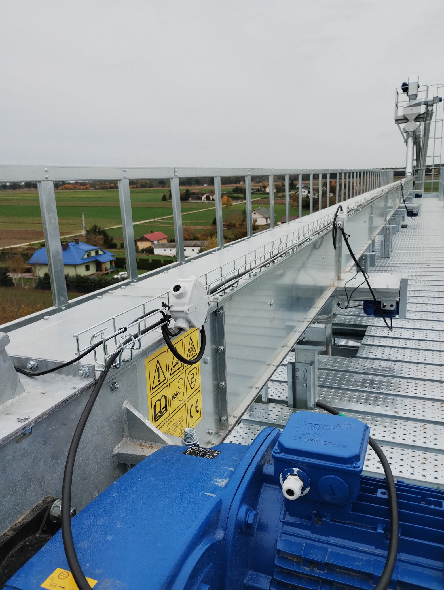 Widok z góry na konstrukcję podtrzymującą instalację elektryczną na zewnątrz budynku w Lublinie, z widocznym niebieskim silnikiem i elementami ostrzegawczymi na żółtych tabliczkach.