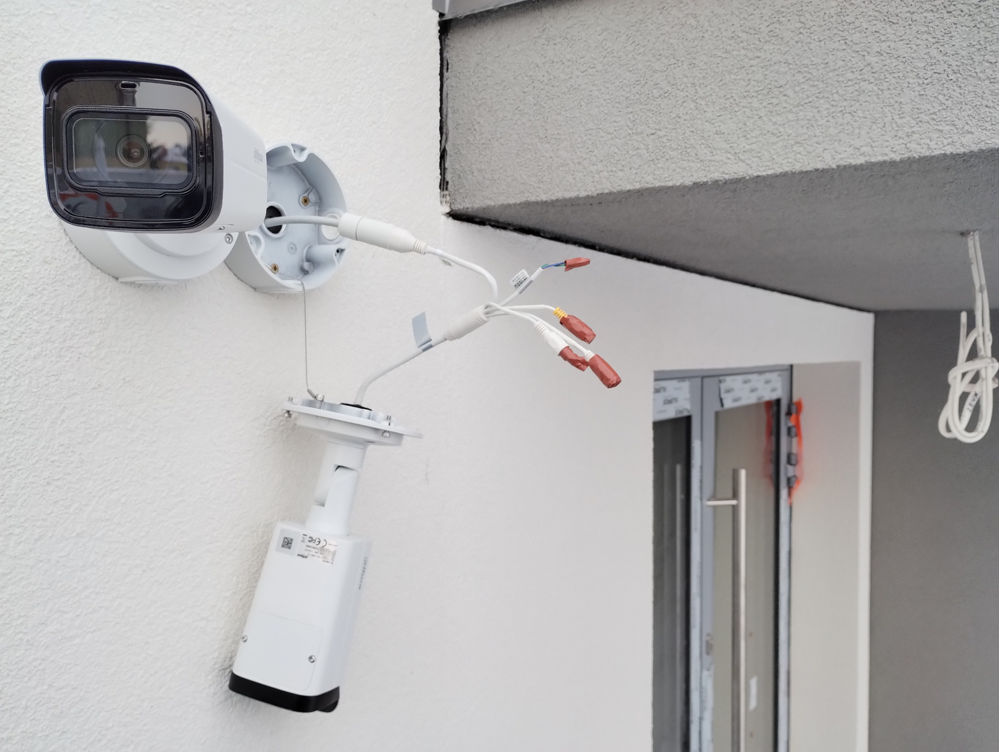 Montaż kamery monitoringu Dahua na elewacji budynku, widoczne kable z końcówkami do podłączenia, uchwyt kamery i korpus kamery z naklejką informacyjną.