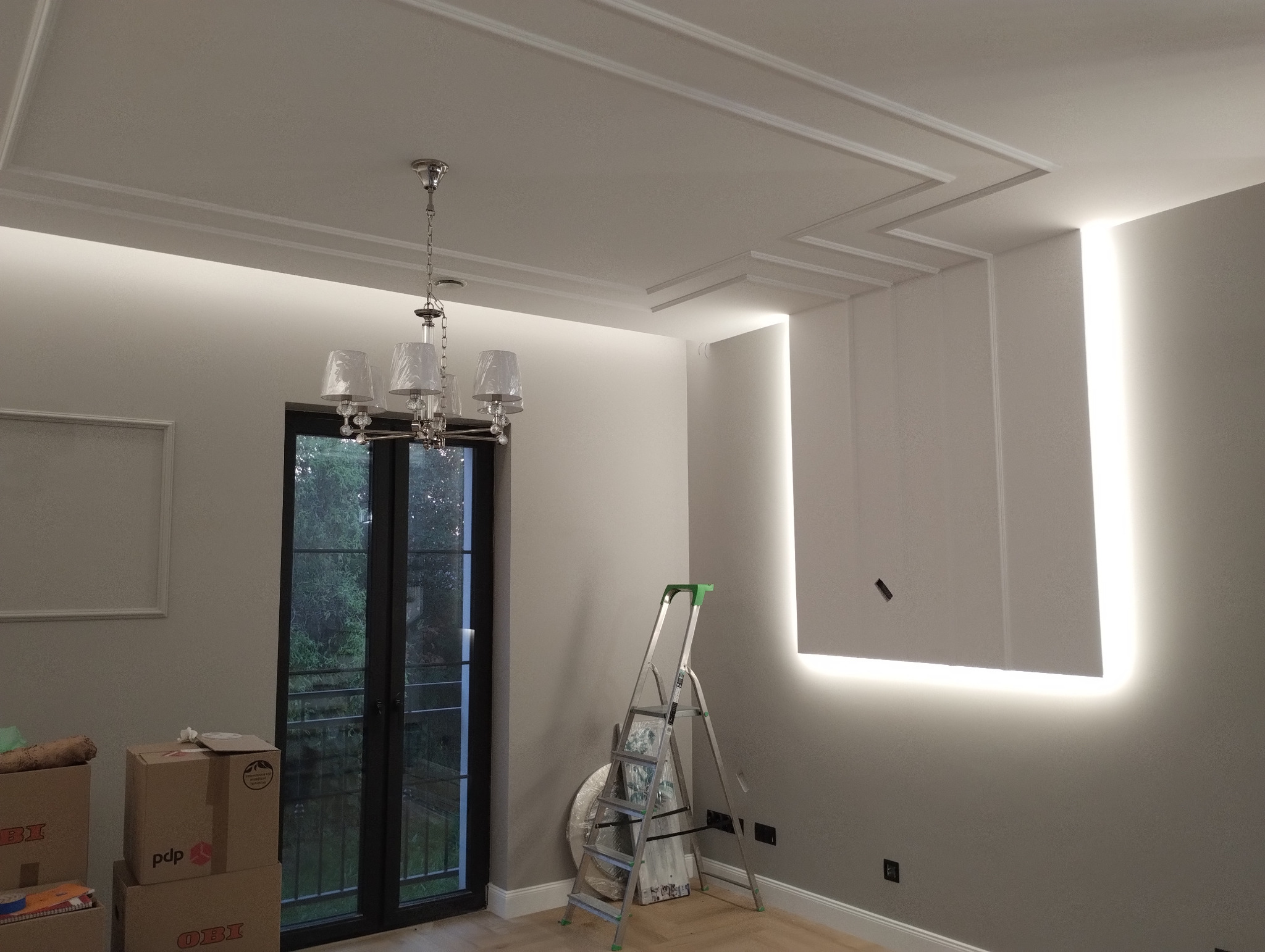 Nowoczesne wnętrze z podświetlanym panelem ściennym, ozdobnym sufitem z listwami i klasycznym żyrandolem; drabina aluminiowa ustawiona przy ścianie.