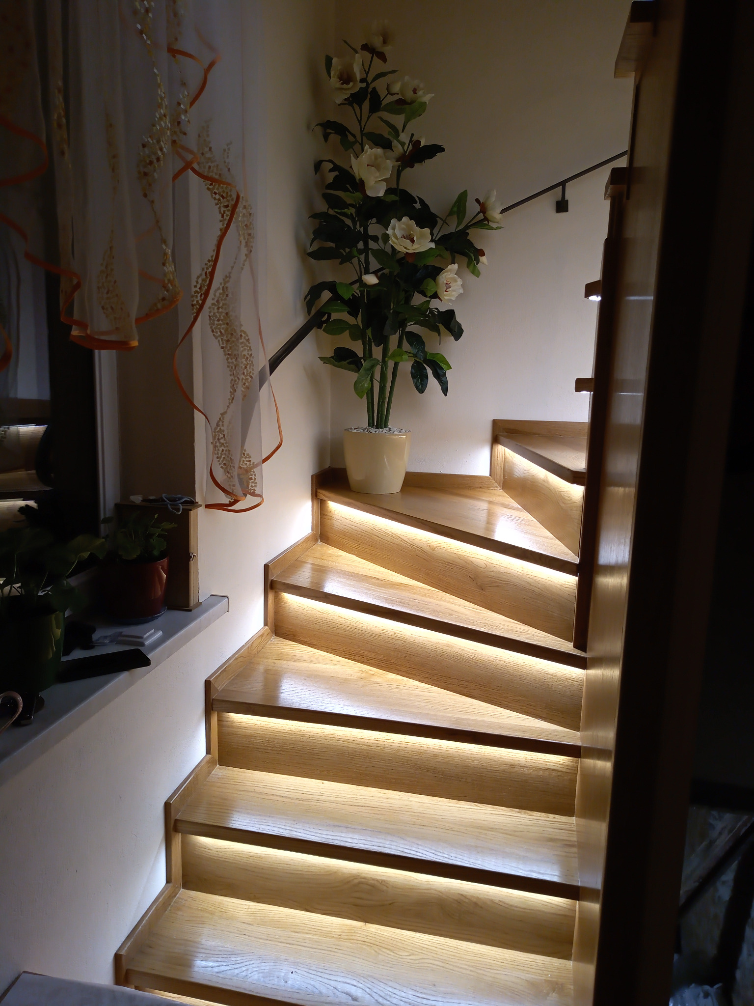 Drewniane schody z podświetleniem LED na każdym stopniu, obok biała ściana z rośliną w doniczce i firanka z pomarańczowymi akcentami.