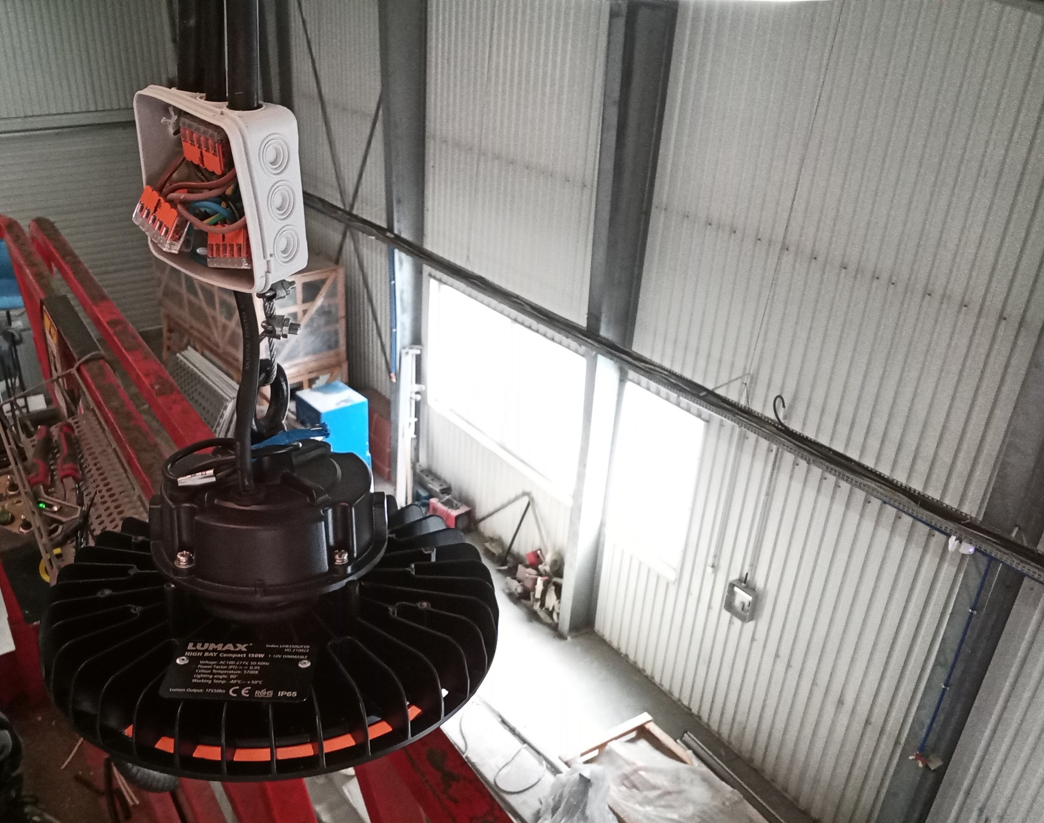 Czarna lampa przemysłowa LUMAX High Bay Compact 150W z pomarańczowym akcentem, zamontowana pod sufitem hali z blachy falistej, widoczna skrzynka elektryczna z podłączonymi przewodami.