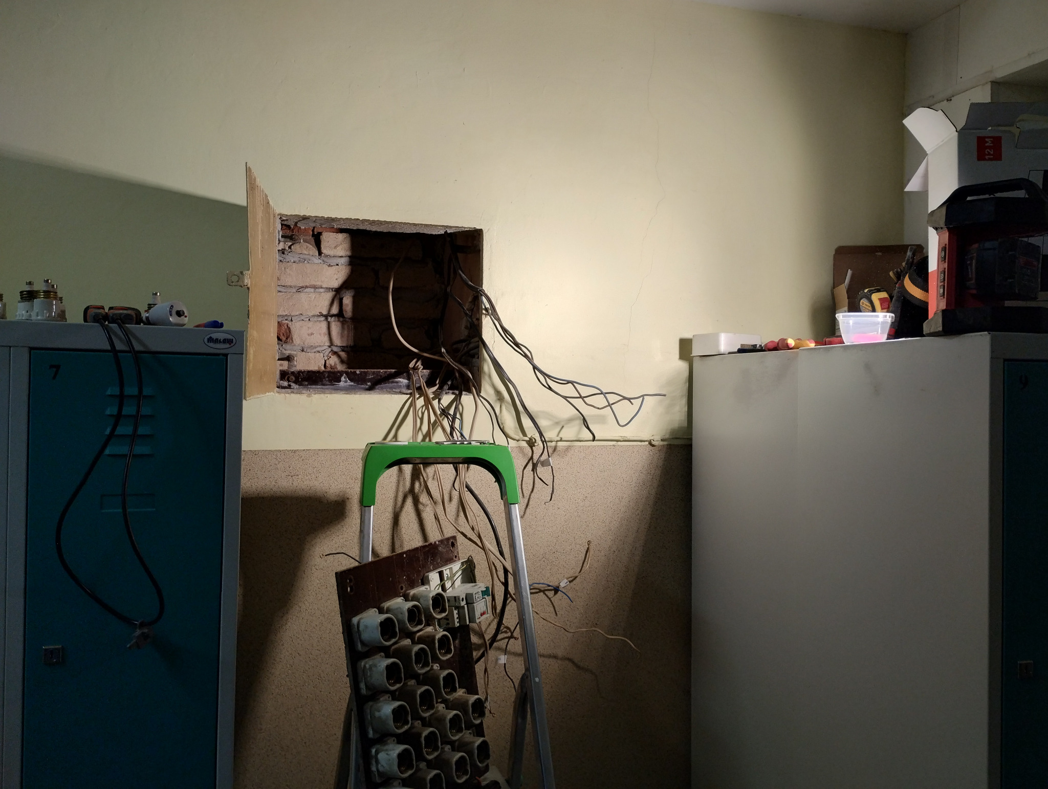 Otwór w ścianie z wystającymi przewodami elektrycznymi, drabina oparta o ścianę z zamontowaną starą tablicą rozdzielczą z ceramicznymi bezpiecznikami, w tle szafki metalowe.