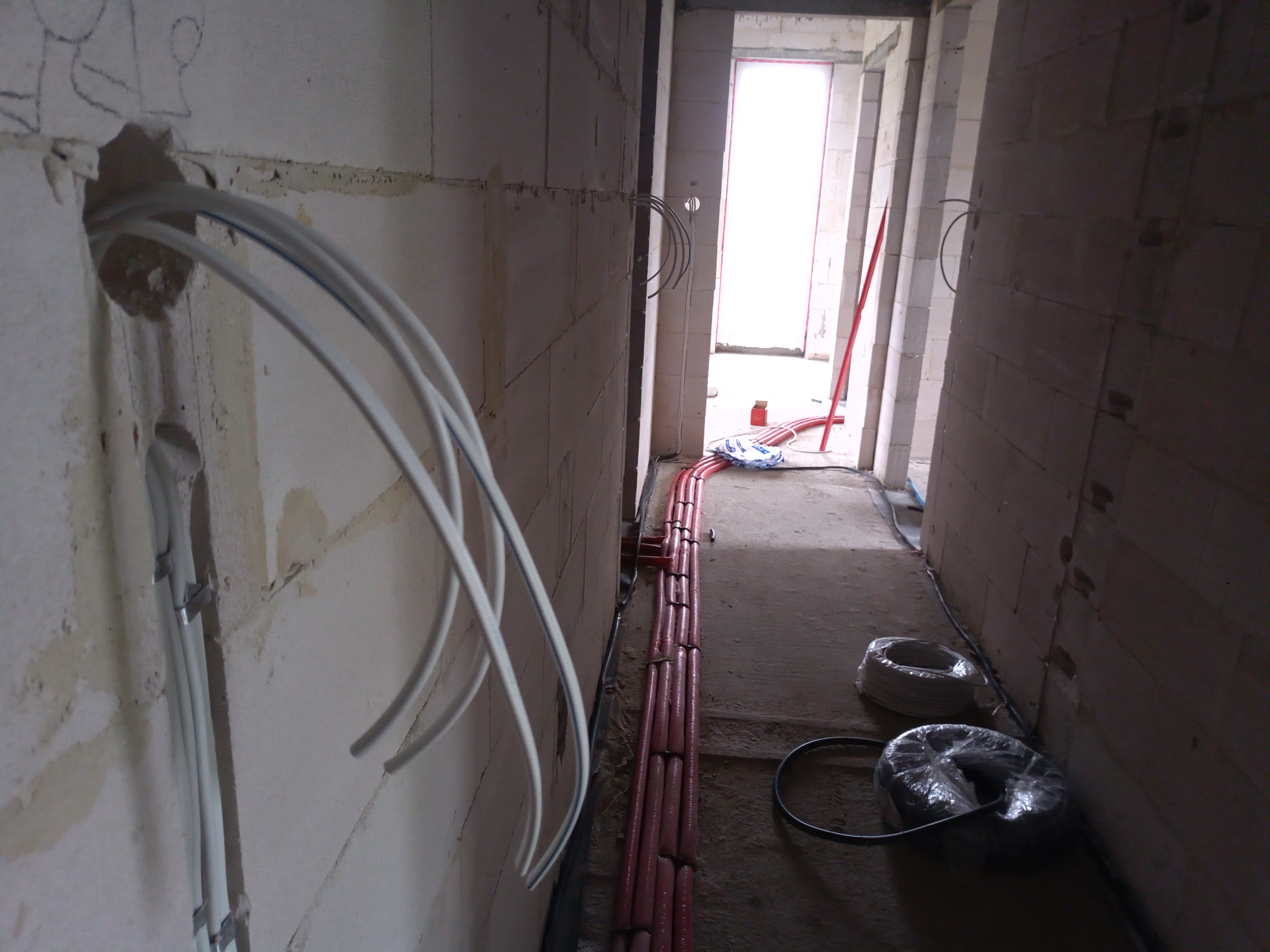 Wnętrze w trakcie prac instalacyjnych: białe kable wychodzące z otworu w surowej ścianie z betonu komórkowego, czerwone rury prowadzone wzdłuż podłogi, w tle zwitki kabli.