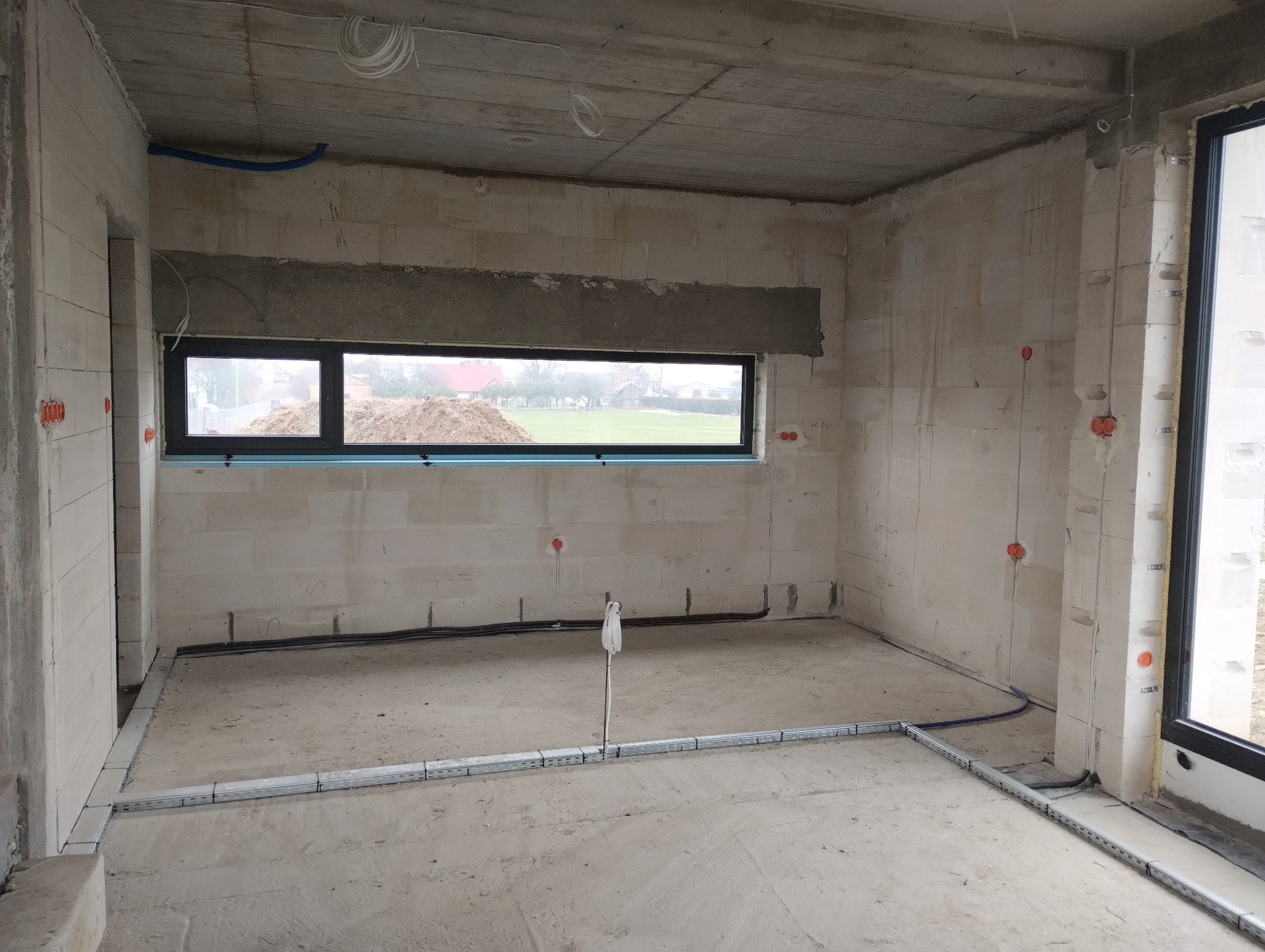 Surowy stan wnętrza budynku w trakcie prac instalacyjnych: widoczne kable elektryczne w peszlach na ścianach i podłodze, puszki instalacyjne w kolorze pomarańczowym, okno z widokiem na zewnątrz.