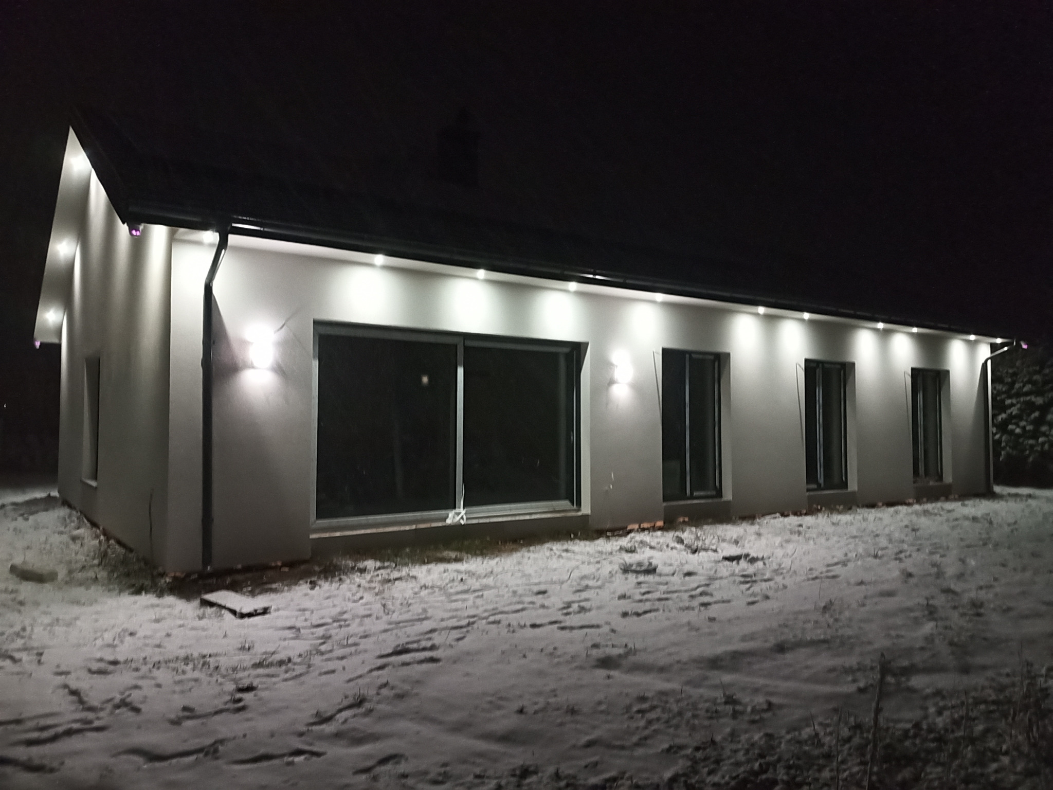 Nowoczesny dom jednorodzinny oświetlony zewnętrznymi lampami LED wzdłuż dachu i na ścianach bocznych, nocna sceneria z pokrywą śnieżną.