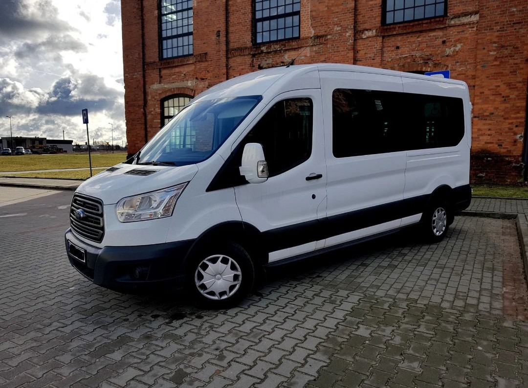 Biały Ford Transit, dziewięcioosobowy bus, zaparkowany na brukowanej nawierzchni przed budynkiem z czerwonej cegły z dużymi oknami. Widok z boku, lekko z przodu.