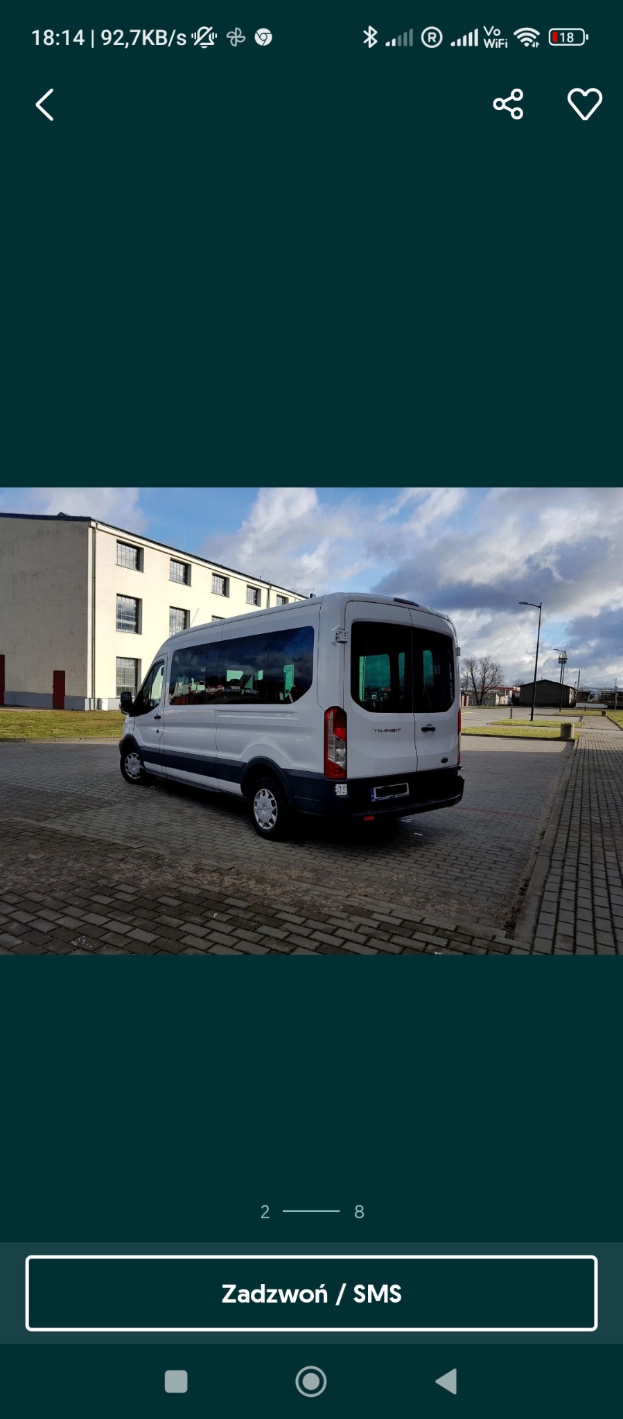 Biały bus marki Ford Transit, zaparkowany na brukowanym placu przed budynkiem z szarymi ścianami i rzędami okien, widok z tyłu i boku.