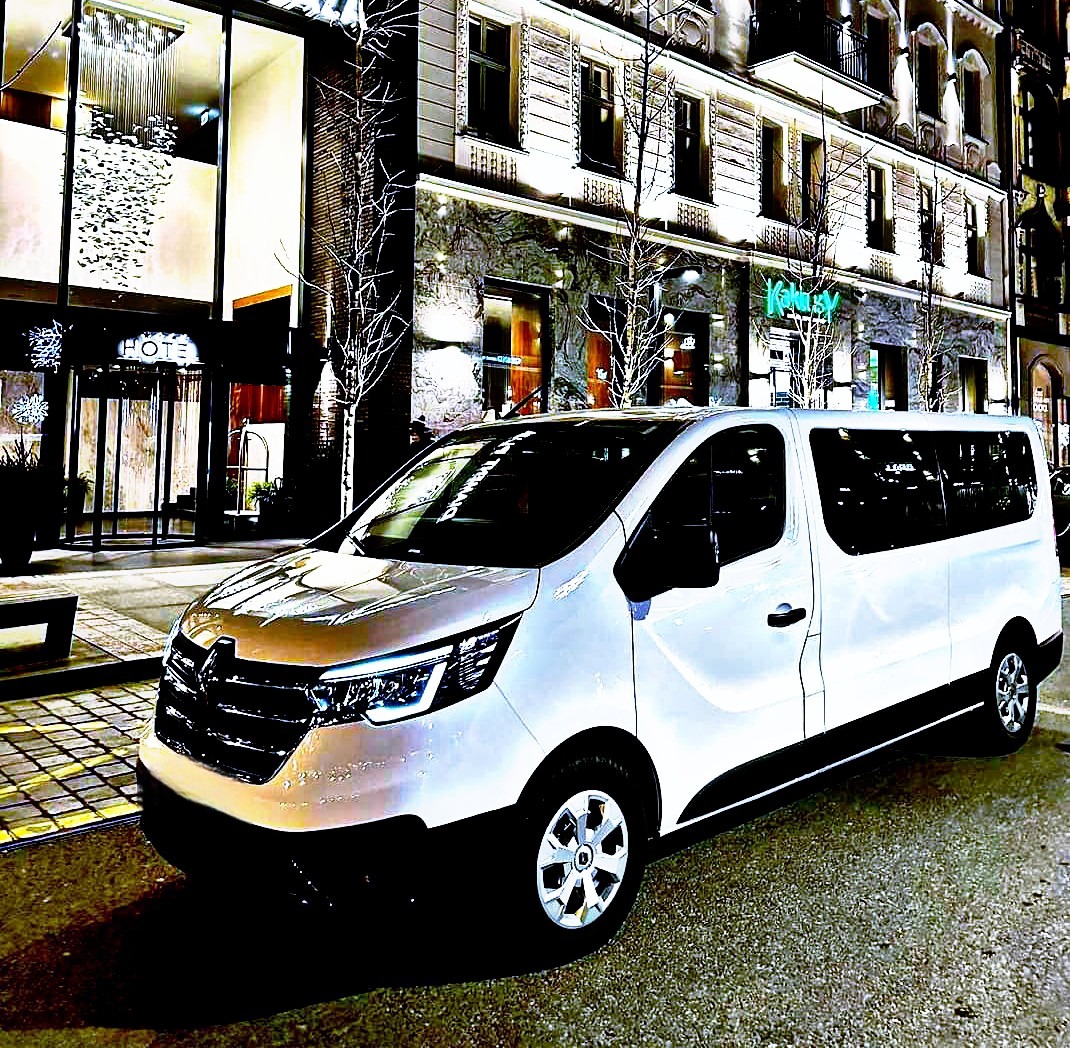 Biały bus Renault Trafic zaparkowany nocą przed eleganckim hotelem z podświetloną fasadą i szklanymi drzwiami wejściowymi.