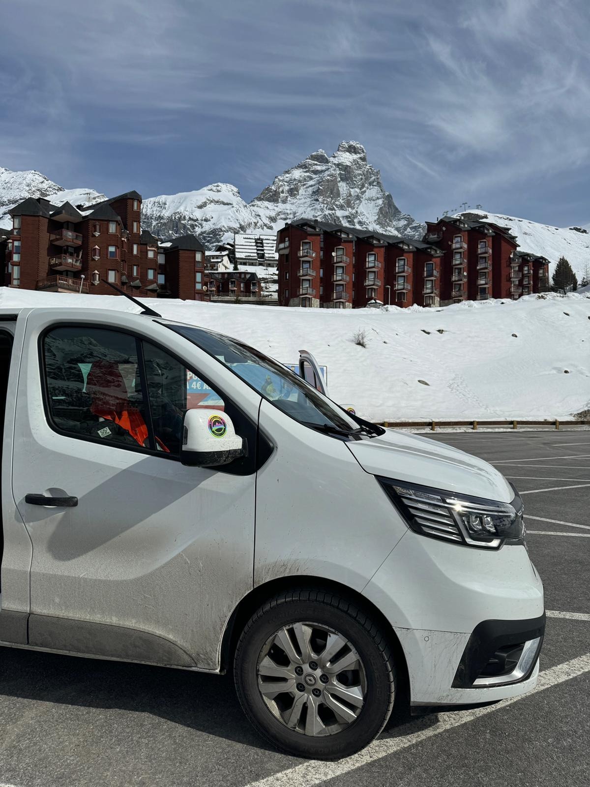 Biały bus na parkingu w zimowej scenerii z widokiem na ośnieżone góry i budynki w tle.