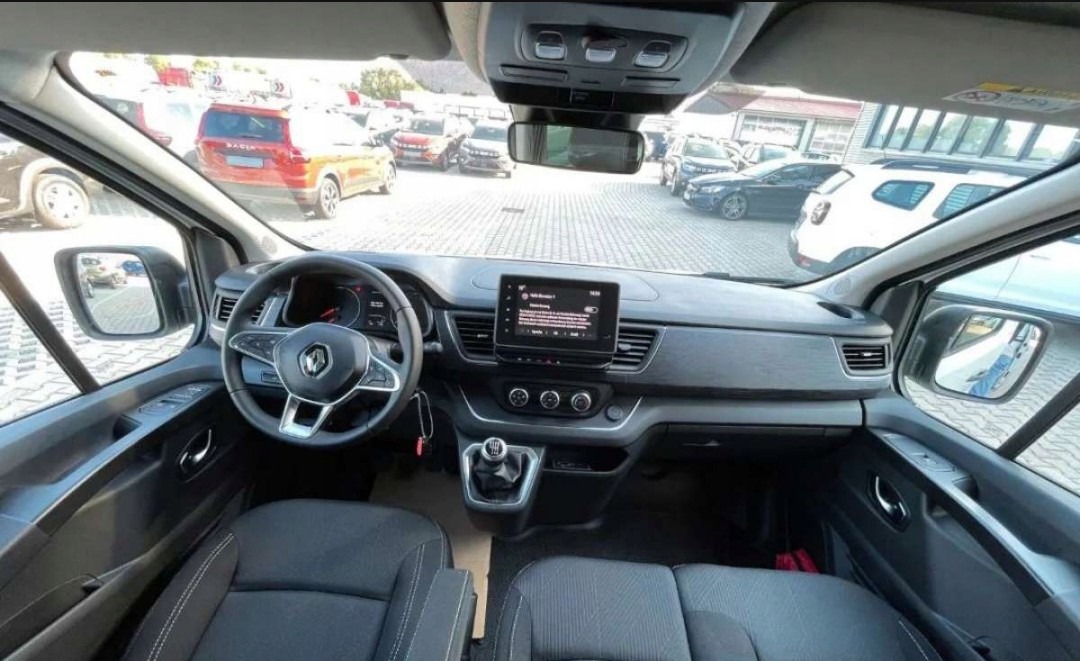 Wnętrze Renault Trafic z widocznym kokpitem, kierownicą, wyświetlaczem i przednimi fotelami. Na zewnątrz widoczne inne zaparkowane pojazdy.