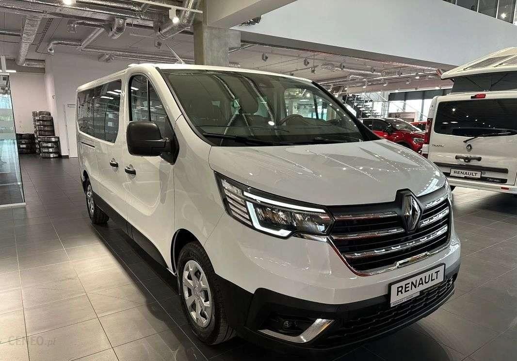 Biały Renault Trafic w salonie samochodowym, widok z przodu i boku, z widocznymi oponami ułożonymi w stos w tle.